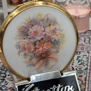 Puede incluir: Un espejo compacto Stratton vintage con marco dorado y diseño floral. El compacto presenta una placa con el nombre "Stratton" en negro y plata. El diseño floral incluye flores rosas, melocotón y moradas.