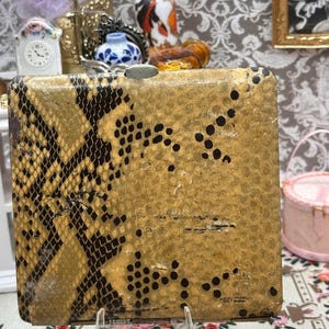 May include: A square cigarette case featuring a textured gold and black snakeskin pattern on the left side and a solid gold surface with scattered black dots on the right. The case has a metallic silver clasp at the top center and a hinged base.