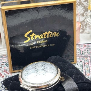 以下が含まれることがあります： シルバーケースと模様のある表面のStrattonコンパクトミラー。黒いベルベットのポーチの上に置かれています。後ろの箱は黒色で、金色の文字で「Stratton England FINE GIFTS SINCE 1860」と書かれています。