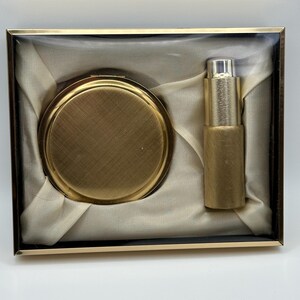 May include: A vintage gold-toned compact and lipstick set presented in a rectangular box. The compact is round with a textured surface, and the lipstick tube is cylindrical. The box has a gold frame and a clear lid, with a cream-colored fabric lining.