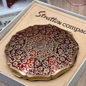May include: A round Stratton compact mirror featuring a deep red enamel finish with intricate gold floral filigree and small blue and red accents. The compact is housed in a beige box with the text Stratton compact and Made in England printed.