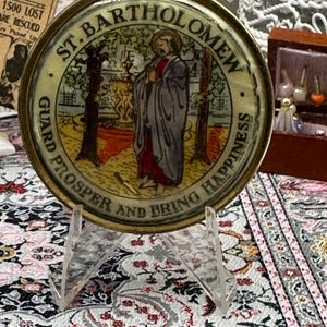Op de afbeelding: Een ronde, antieke sieradendoos met een goudkleurige rand. Het midden toont een illustratie van St. Bartholomew, met de tekst "GUARD PROSPER AND BRING HAPPINESS" en "ST. BARTHOLOMEW". De doos wordt getoond op een heldere acrylstandaard.