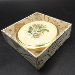 Puede incluir: Un compacto redondo de color crema con un borde dorado, con un diseño floral y un colibrí. Se presenta en una caja decorativa con letras doradas y detalles de estrellas. La caja dice "London".