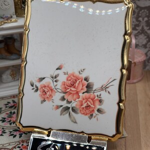 May include: A rectangular Stratton compact mirror with a white enamel surface featuring a floral design of three coral-colored roses with green leaves. The compact has a scalloped gold-tone metal frame and a black rectangular base with the word Stratton.
