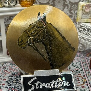 May include: A vintage Stratton compact mirror with a gold finish. The compact features an embossed horse head design and a black and silver nameplate that reads "Stratton".