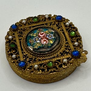Puede incluir: Una caja de baratijas redonda y dorada de estilo vintage, con intrincados detalles de filigrana. La caja presenta un diseño floral ovalado pintado a mano en el centro, rodeado de acentos azules, verdes y perlados. Tiene una tapa con bisagras y un cierre decorativo.