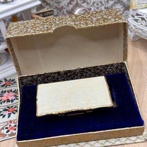 以下が含まれることがあります： 金色の縁取りが施されたクリーム色の質感のある長方形のケースが、開いたヒンジ付きの箱の中に収められています。箱の外側には金と白の花柄があしらわれ、内側には深い青色のベルベットの裏地が敷かれています。