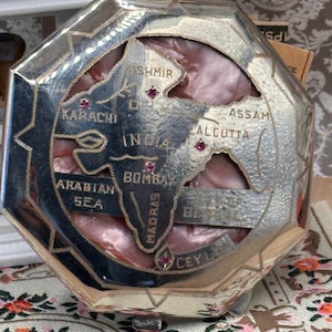Peut inclure: Une boîte octogonale en métal argenté avec une carte découpée de l'Inde sur le couvercle. La carte présente des inscriptions gravées pour Kashmir, Karachi, India, Bombay, Madras, Assam, Calcutta, Arabian Sea et Ceylon, avec des pierres rouges.