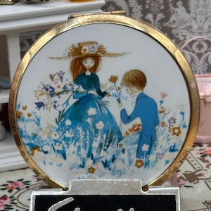 May include: A round gold-tone metal compact mirror featuring a white enamel lid with a colorful illustration of a person in a blue dress and a person in a blue suit standing in a field of flowers. A black rectangular plaque with the word Stratton.