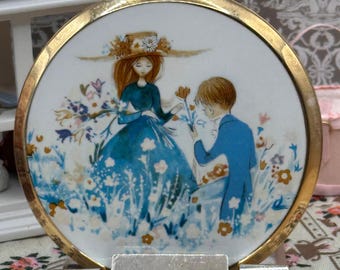 May include: A round gold-tone metal compact mirror featuring a white enamel lid with a colorful illustration of a person in a blue dress and a person in a blue suit standing in a field of flowers. A black rectangular plaque with the word Stratton.