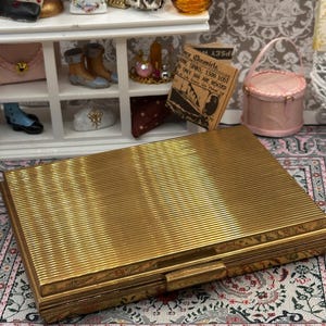 May include: A gold-coloured, rectangular box with a ribbed surface and a clasp. The box is set on a patterned rug, with miniature decorative items in the background, including a clock, shoes, and a small pink purse.