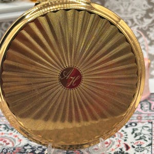 May include: A gold-coloured compact mirror with a sunburst pattern. The compact is circular and has a textured surface. The initials "AH" are engraved in a circular emblem in the centre of the compact.