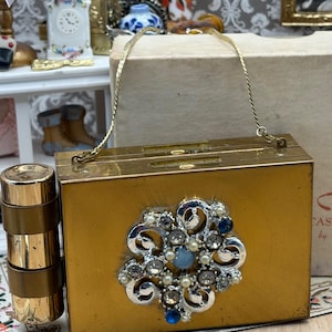 May include: A rectangular gold-toned metal clutch purse featuring a thin gold chain strap and a prominent silver-toned floral brooch ornament set with clear, blue, and white stones. A matching gold-toned cylindrical lipstick case is attached to the side.