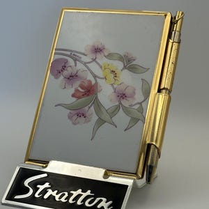 May include: A vintage Stratton compact mirror with a gold frame and a floral design. The compact features a black and silver Stratton nameplate. The pen is attached to the side of the compact.