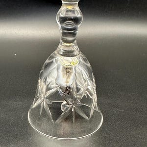 Puede incluir: Campana de cristal transparente con un diseño de estrella en la parte inferior. La campana tiene una base redondeada y un mango largo y delgado. El cristal transparente permite el paso de la luz y es probablemente un objeto decorativo.