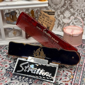 May include: A vintage red comb with a black and silver Stratton compact. The comb is made of a translucent red material. The compact is black with a silver nameplate that reads "Stratton". The compact has a gold emblem of two people.