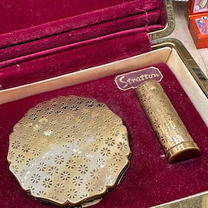 May include: A vintage Stratton vanity set featuring a round gold-tone powder compact and a matching lipstick case, both decorated with a repeating floral pattern. The items are presented in a rectangular case lined with deep red velvet fabric.
