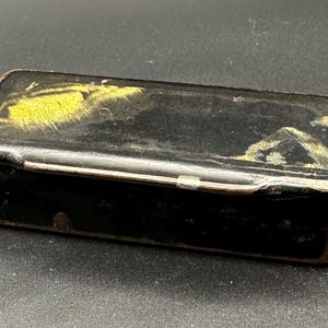 May include: Rectangular black box with a hinged lid. The box has a worn appearance with gold and yellow accents on the top. The edges of the box are a lighter brown colour. The box is closed with a small metal clasp.