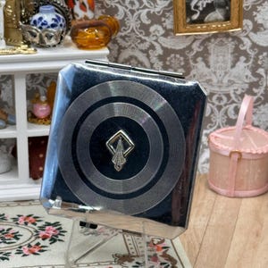 May include: A square silver-toned metal compact case featuring a central black circular inlay with concentric etched rings and a small, diamond-shaped silver emblem at the center. The compact has a polished, reflective finish and a hinged metal closure.