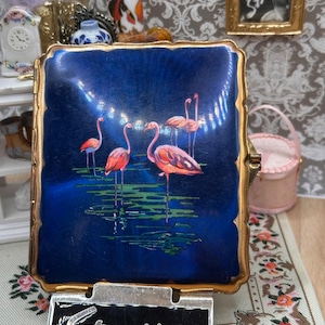 May include: A square Stratton compact with a deep blue enamel finish featuring a painted scene of four pink flamingos standing in green water. The compact has a gold-toned metal border and is displayed with a black plaque bearing the name Stratton.