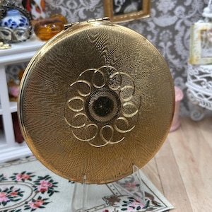 May include: A round, gold-toned metal compact mirror featuring a textured, cross-hatched surface finish. The center of the lid is decorated with an engraved circular pattern of interconnected loops surrounding a small, solid, dark-colored center circle.