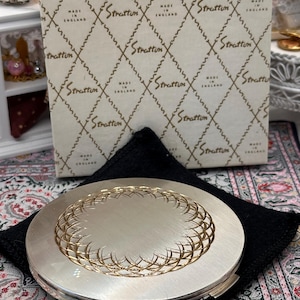 May include: A round silver-tone Stratton compact mirror featuring a central gold-tone circular lattice pattern. The compact is presented on a black fabric pouch in front of its original off-white box, which is printed with a repeating Stratton logo.