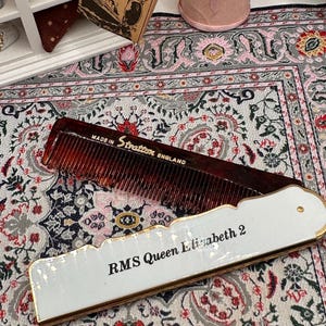 May include: A vintage brown comb with the text "MADE IN Stratton ENGLAND" and a white rectangular item with "RMS Queen Elizabeth 2" printed on it, with gold trim. The items are on a patterned rug.