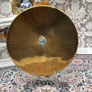 Pode incluir: Um espelho compacto redondo dourado com um design radial. O centro apresenta uma pequena pedra preciosa azul clara rodeada por uma moldura prateada. O compacto é exibido em um suporte transparente.