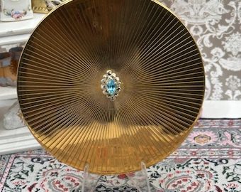 May include: A gold-colored, round compact mirror with a radial design. The center features a small, light blue gemstone surrounded by a silver-colored setting. The compact is displayed on a clear stand, showcasing its decorative appeal.