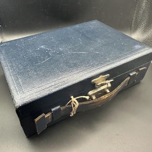 Puede incluir: Un maletín de cuero azul vintage con cierre metálico y asa de cuero. El maletín rectangular muestra signos de desgaste, lo que sugiere que es una antigüedad o un artículo muy usado. El asa está sujeta con herrajes metálicos.