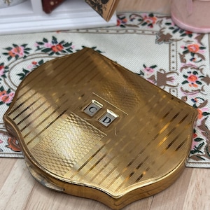 May include: A gold-toned metal compact case with a scalloped edge and a textured, striped surface. The center features a small rectangular plaque with the letters C and D in a serif font. The compact has a hinged closure and a polished finish.