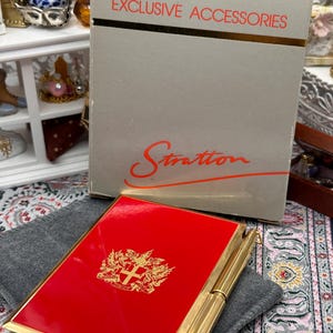 May include: A red Stratton compact with gold trim and a gold crest design. The compact is next to a gray box with the words "EXCLUSIVE ACCESSORIES" and the Stratton logo. The compact is on a gray cloth.