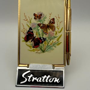 May include: A vintage Stratton compact mirror with a gold-tone frame. The compact features a painted scene of butterflies and thistles. The name "Stratton" is displayed on a silver and black nameplate.