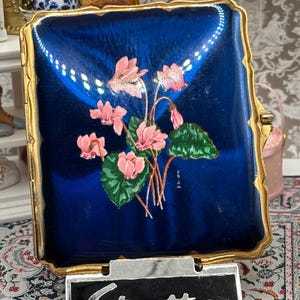 May include: A vintage Stratton compact mirror with a blue enamel finish and a floral design. The compact features a gold-tone frame and a black and silver Stratton nameplate. The floral design includes pink flowers and green leaves.