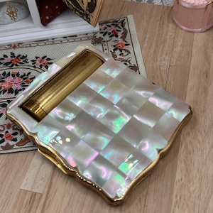 May include: A square compact mirror featuring a grid of iridescent mother-of-pearl tiles on the lid. The compact has a gold-toned metal frame and a textured gold-toned cylindrical hinge mechanism along one side, with a scalloped edge design.