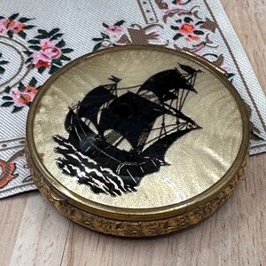 May include: A round gold-toned metal compact mirror featuring a black silhouette of a sailing ship on the lid. The lid has a textured, wavy engraved pattern, and the side of the compact is decorated with an intricate floral relief design.