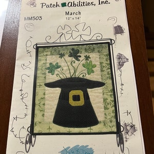 May include: A paper craft pattern by Patch Abilities, Inc. featuring a leprechaun hat and shamrocks. The design is framed with a black border and includes the text "March" and dimensions of 12" x 14".
