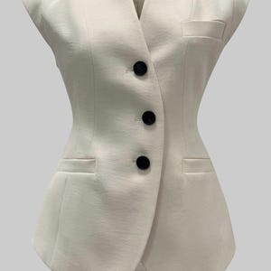 May include: Ivory-colored sleeveless vest with a tailored design. Features a V-neck, three black buttons, and two front pockets. The vest has a fitted silhouette and a classic, elegant style.