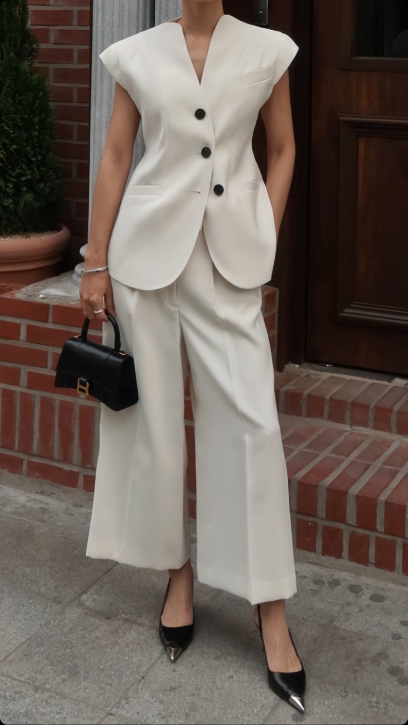 May include: A cream-colored sleeveless vest with three black buttons, paired with matching wide-leg pants. A black handbag and black pointed-toe heels complete the outfit. The ensemble is a modern take on a classic suit.