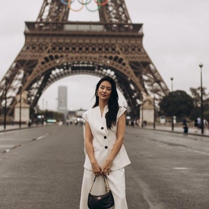 May include: A woman in a white suit and black purse stands in front of the Eiffel Tower. The suit includes a sleeveless blazer and wide-leg pants. The black purse has a gold chain strap. The Eiffel Tower is in the background.