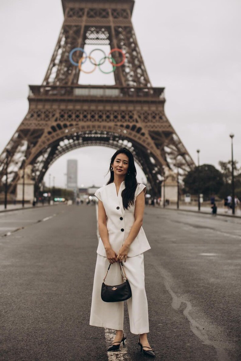 May include: A woman in a white suit and black purse stands in front of the Eiffel Tower. The suit includes a sleeveless blazer and wide-leg pants. The black purse has a gold chain strap. The Eiffel Tower is in the background.