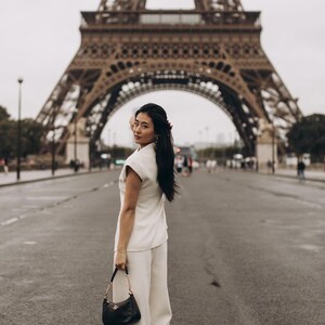 May include: A woman in a cream-colored pantsuit and black handbag stands on a wet street in front of the Eiffel Tower. The black handbag has a gold chain strap. The Eiffel Tower is in the background.
