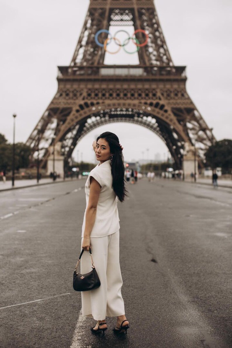 May include: A woman in a cream-colored pantsuit and black handbag stands on a wet street in front of the Eiffel Tower. The black handbag has a gold chain strap. The Eiffel Tower is in the background.