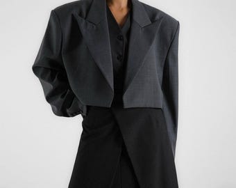 May include: A cropped grey blazer with a wide lapel and large shoulder pads. The blazer is made of a textured fabric and has a slightly oversized fit.