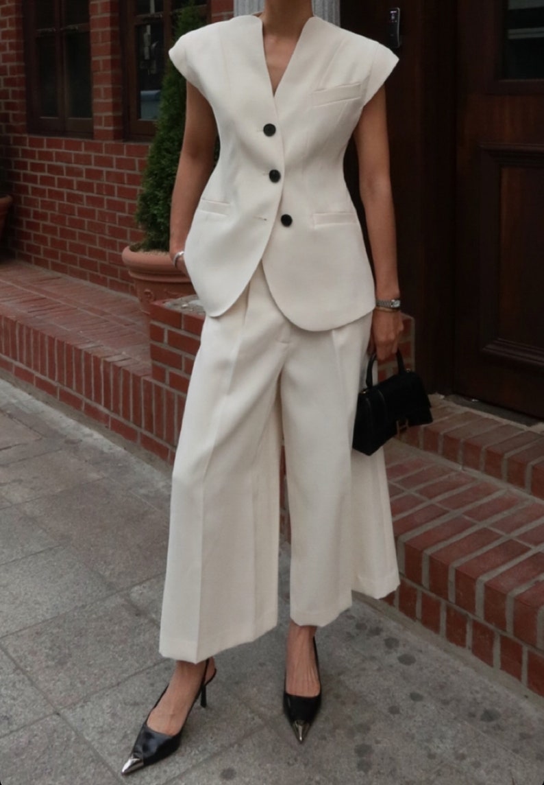 May include: Cream-colored women's suit featuring a sleeveless blazer with black buttons and wide-leg pants. The outfit is paired with black heels and a small black handbag. The suit is made of a light fabric, suitable for warmer weather.