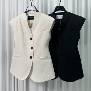 May include: Two sleeveless vests hanging on a rack. One is cream-colored, the other is black. Both have a V-neck, three buttons, and a small pocket. The vests are tailored with a structured design.