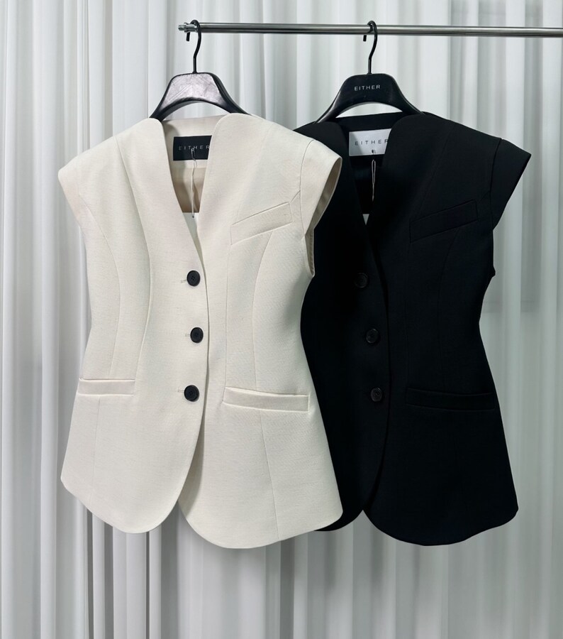 May include: Two sleeveless vests hanging on a rack. One is cream-colored, the other is black. Both have a V-neck, three buttons, and a small pocket. The vests are tailored with a structured design.