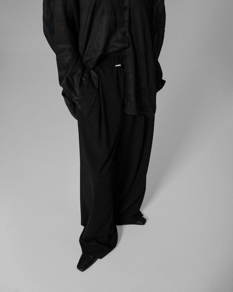 Pu&ograve; includere: Una camicia nera a maniche lunghe con chiusura a bottoni e un paio di pantaloni neri a gamba larga.