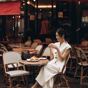 May include: A woman in a white outfit sits at a cafe table, holding a cup. A croissant sits on a plate. The cafe has round tables, wicker chairs, and a red awning. The scene evokes a European cafe setting.