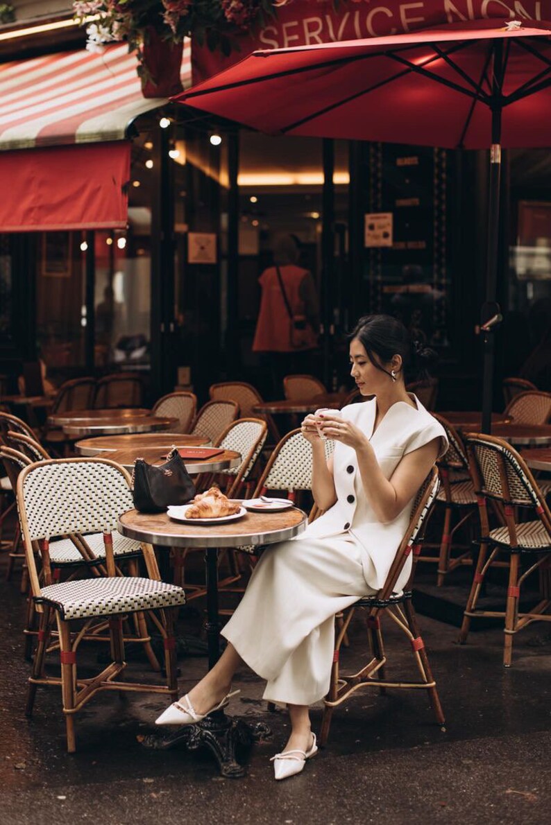 May include: A woman in a white outfit sits at a cafe table, holding a cup. A croissant sits on a plate. The cafe has round tables, wicker chairs, and a red awning. The scene evokes a European cafe setting.